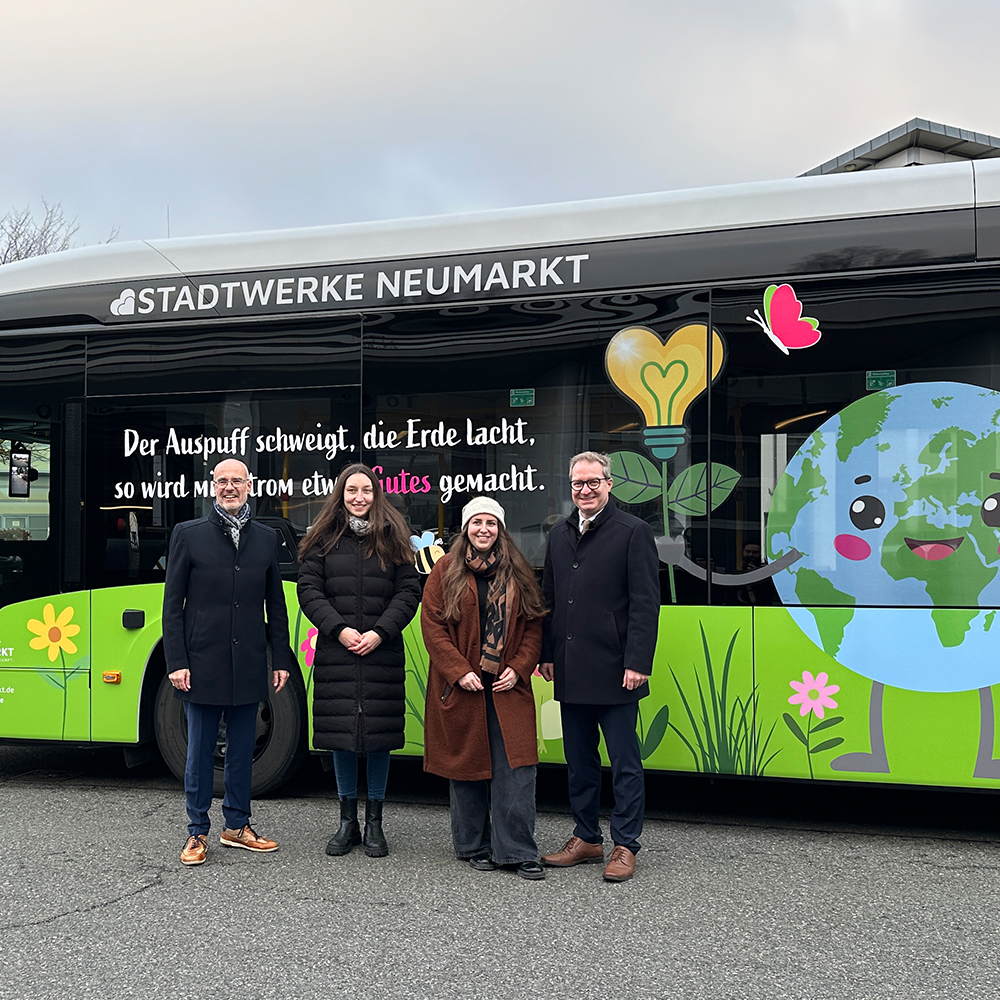 Vier Personen stehen nebeneinander vor einem grünen Stadtbus der Stadtwerke Neumarkt. Der Bus ist mit bunten Illustrationen zum Thema Umwelt und Nachhaltigkeit gestaltet, darunter eine lachende Erde und Pflanzen. Auf dem Bus steht der Schriftzug ‚Der Auspuff schweigt, die Erde lacht‘.