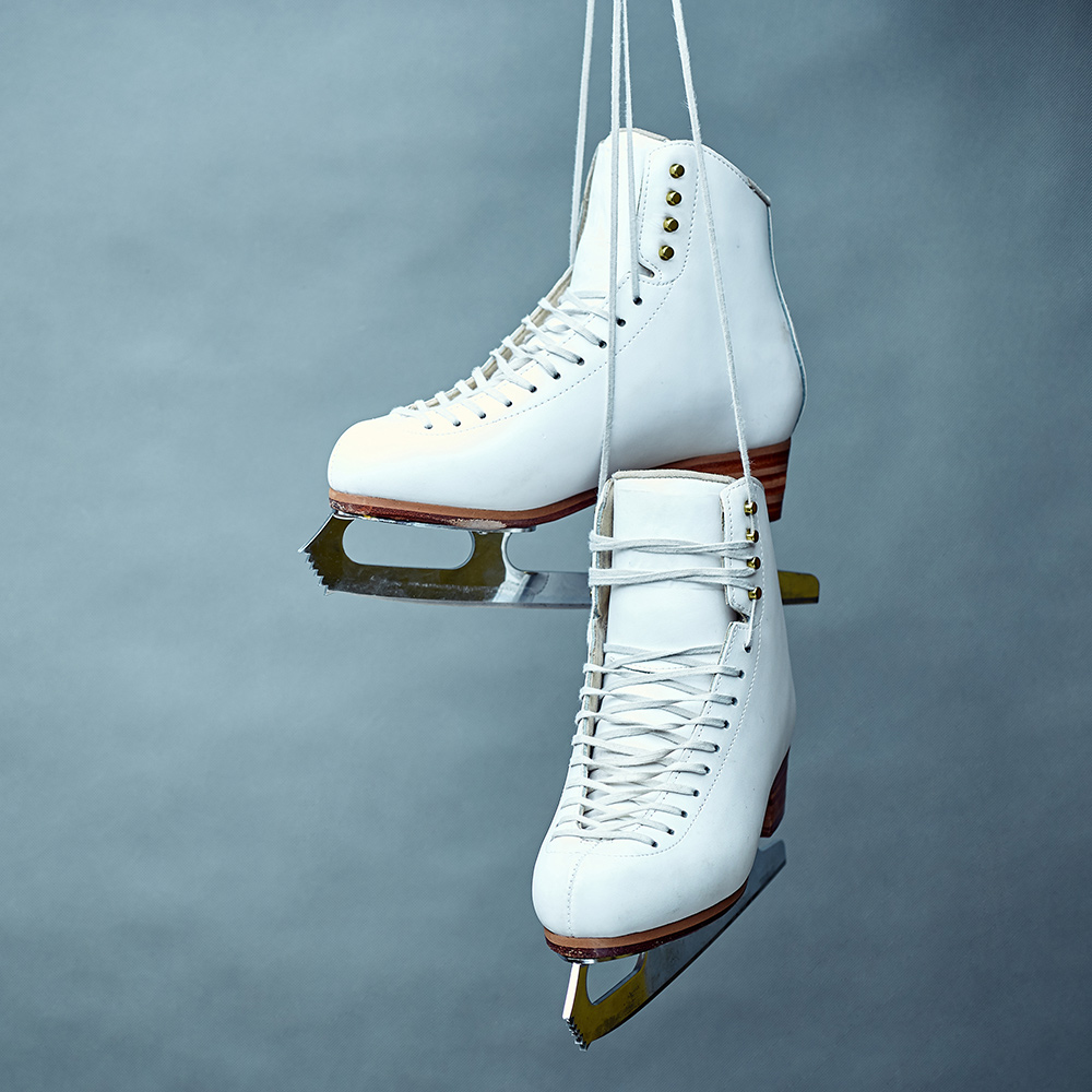 Ein Paar weiße Eiskunstlauf-Schlittschuhe hängt an ihren langen, weißen Schnürsenkeln vor einem gleichmäßig graublauen Hintergrund. Die Kufen glänzen leicht im Licht.